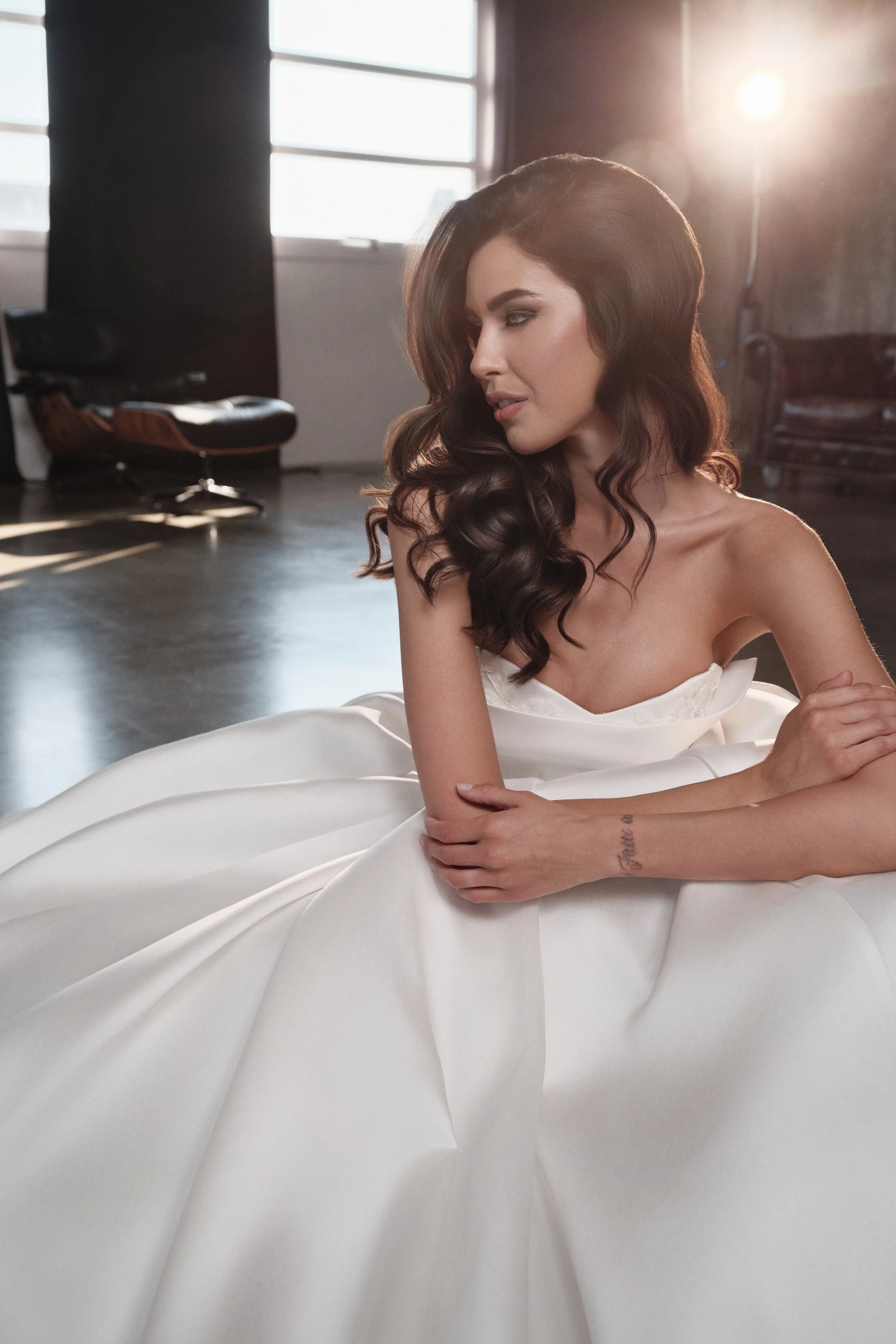 A woman with long wavy dark hair in a white dress sitting on a white surface in a studio with large windows and sunlight streaming in.