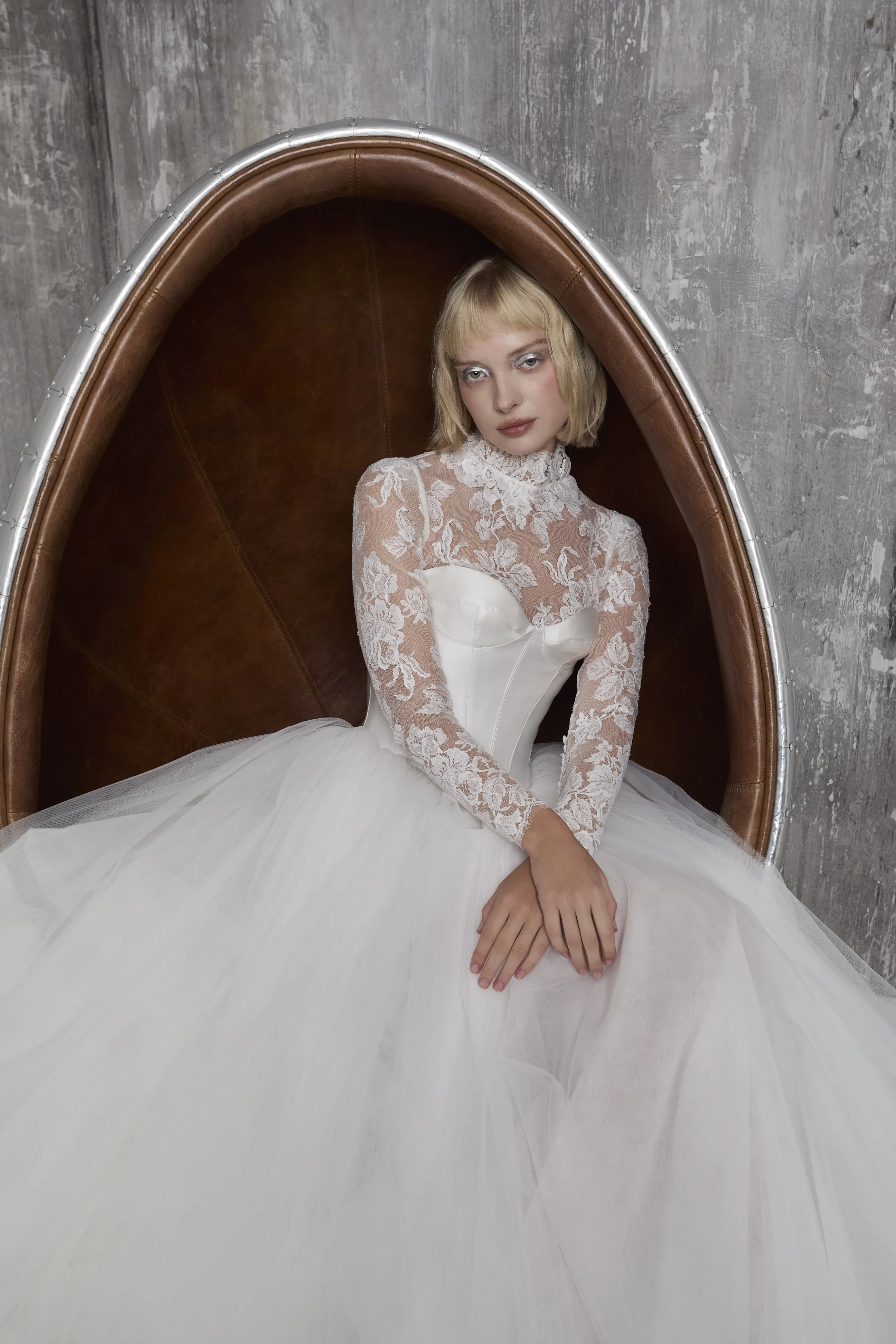 A woman in a white lace long sleeve top and a white tulle skirt sitting in a brown leather and metal chair with a concrete wall in the background.