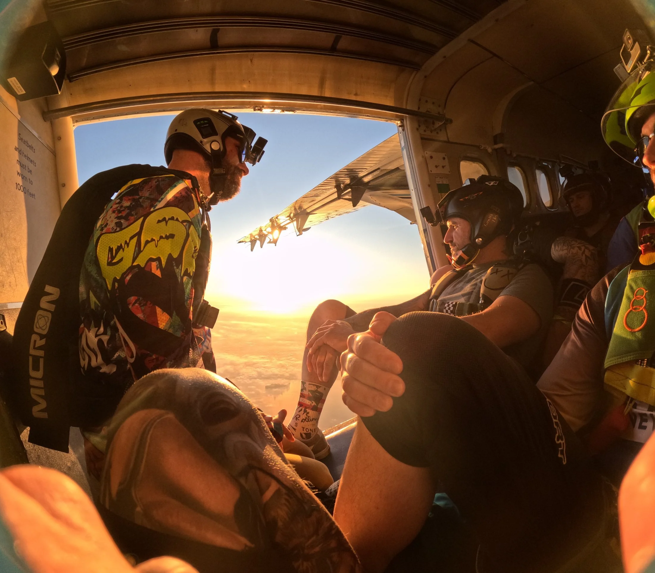 View from inside a helicopter with several people wearing helmets and headlamps. One person is handing something to another. The sunset sky is visible through the open helicopter door.