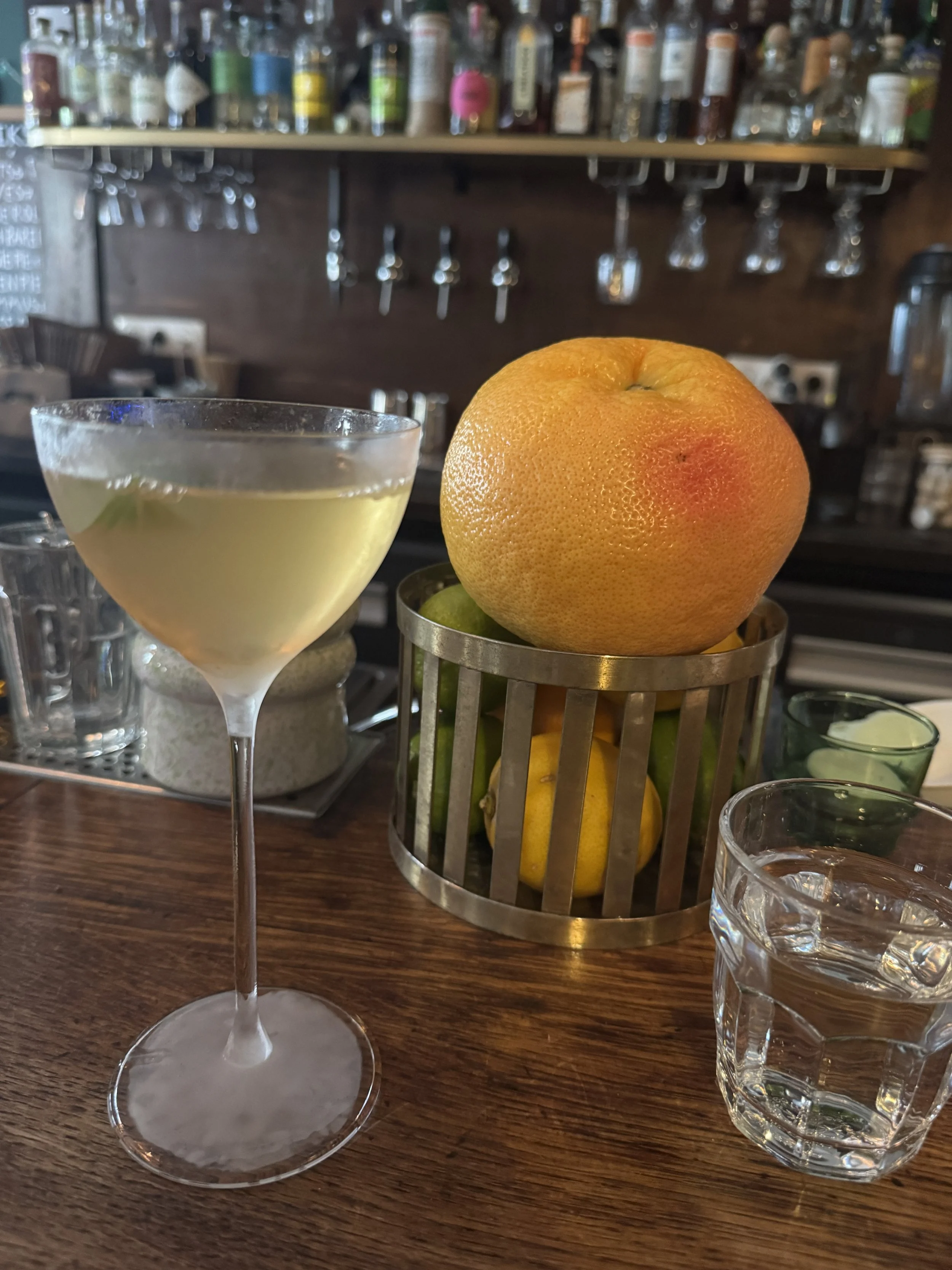 A cocktail glass filled with a light-colored drink, a large grapefruit, a small lime, and a glass of water on a wooden bar counter with bottles and glasses in the background.