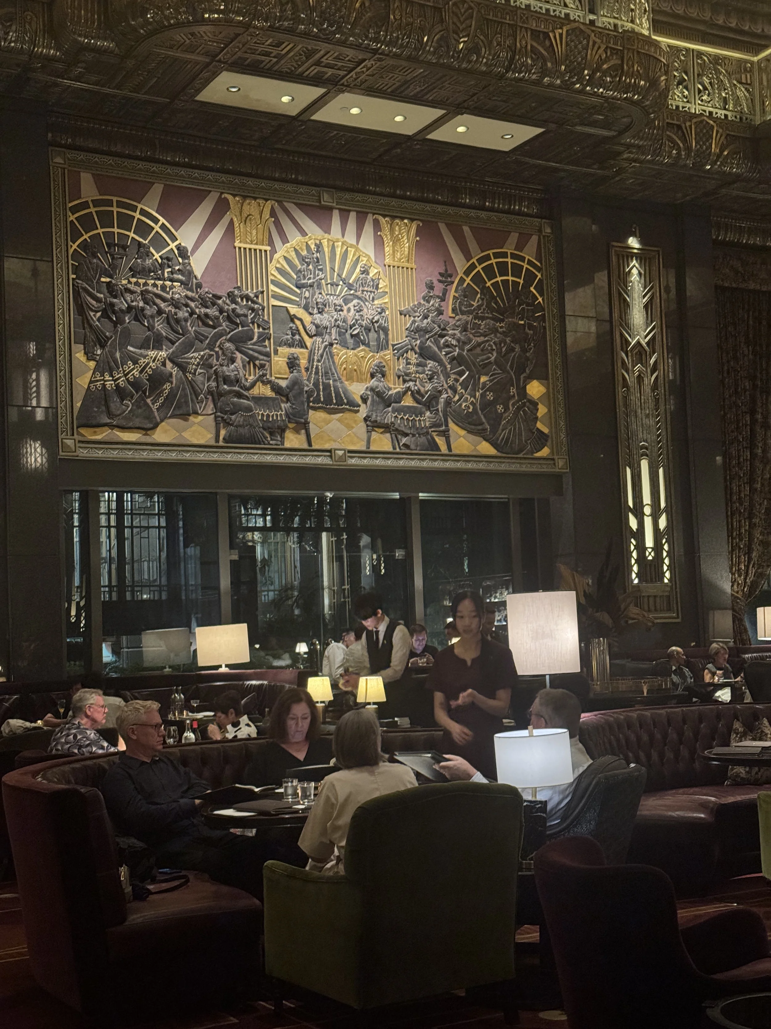 A bustling restaurant with elegant decor, including a large, ornate wall art piece depicting a traditional scene with musicians and dancers. Diners are seated at tables, and staff are attending to the guests.