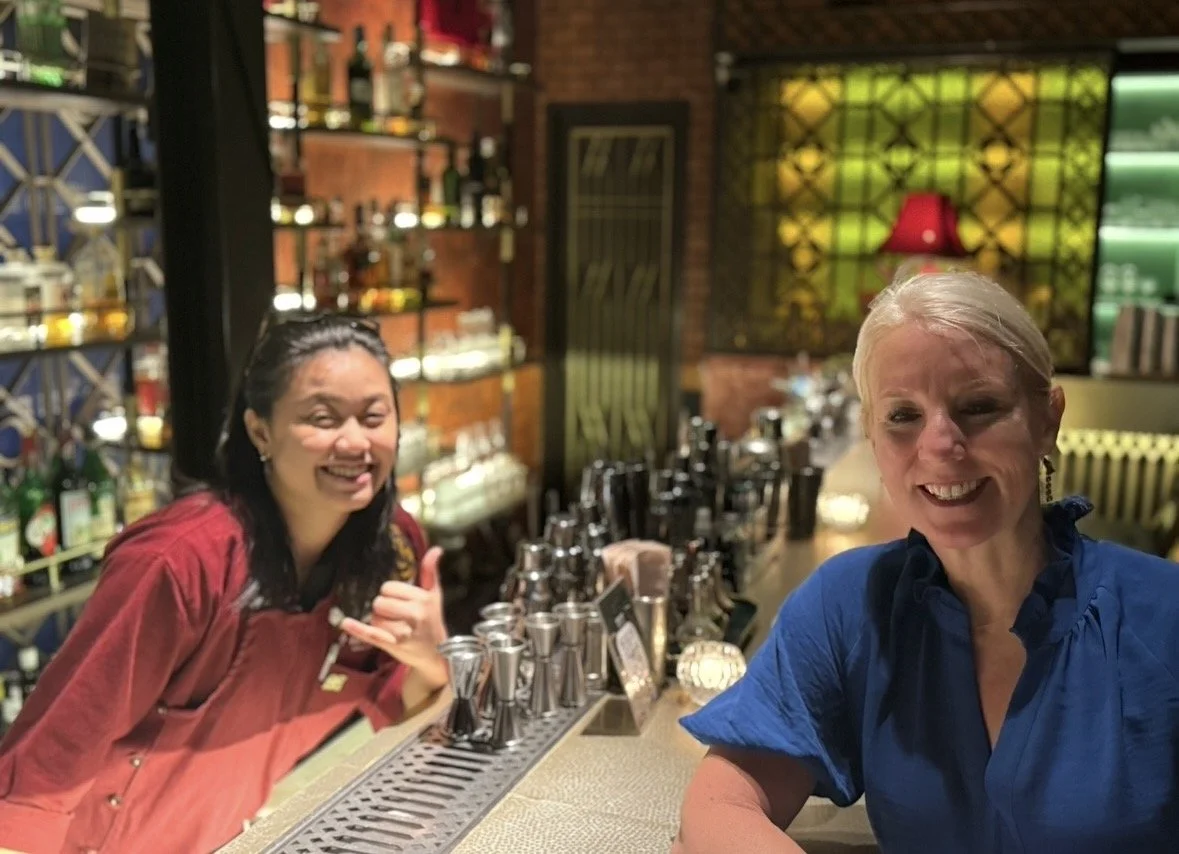 Two women smiling at a bar with various bottles of alcohol in the background.