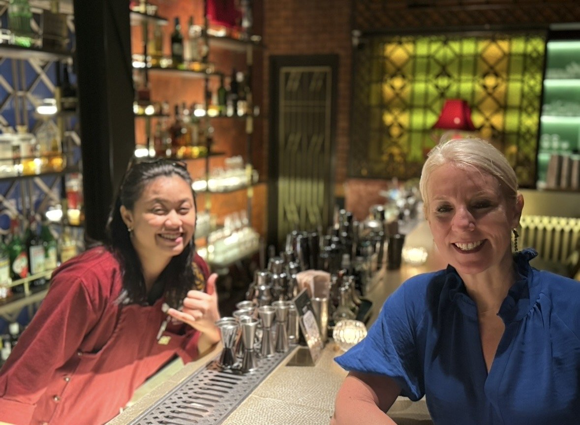 Two women smiling behind a bar counter in a cozy, dimly lit bar or restaurant. Shelves with bottles and glasses are visible in the background.