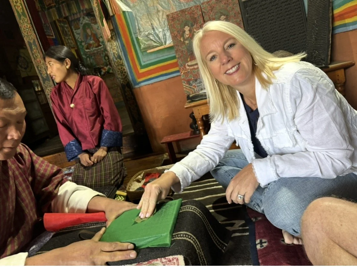 A woman with blonde hair smiling while sitting on the floor and touching a green book or box held by a man. Two other people are visible in the background, one wearing traditional clothing and standing, the other partially visible. The setting appears to be indoors with colorful, traditional decor on the walls.