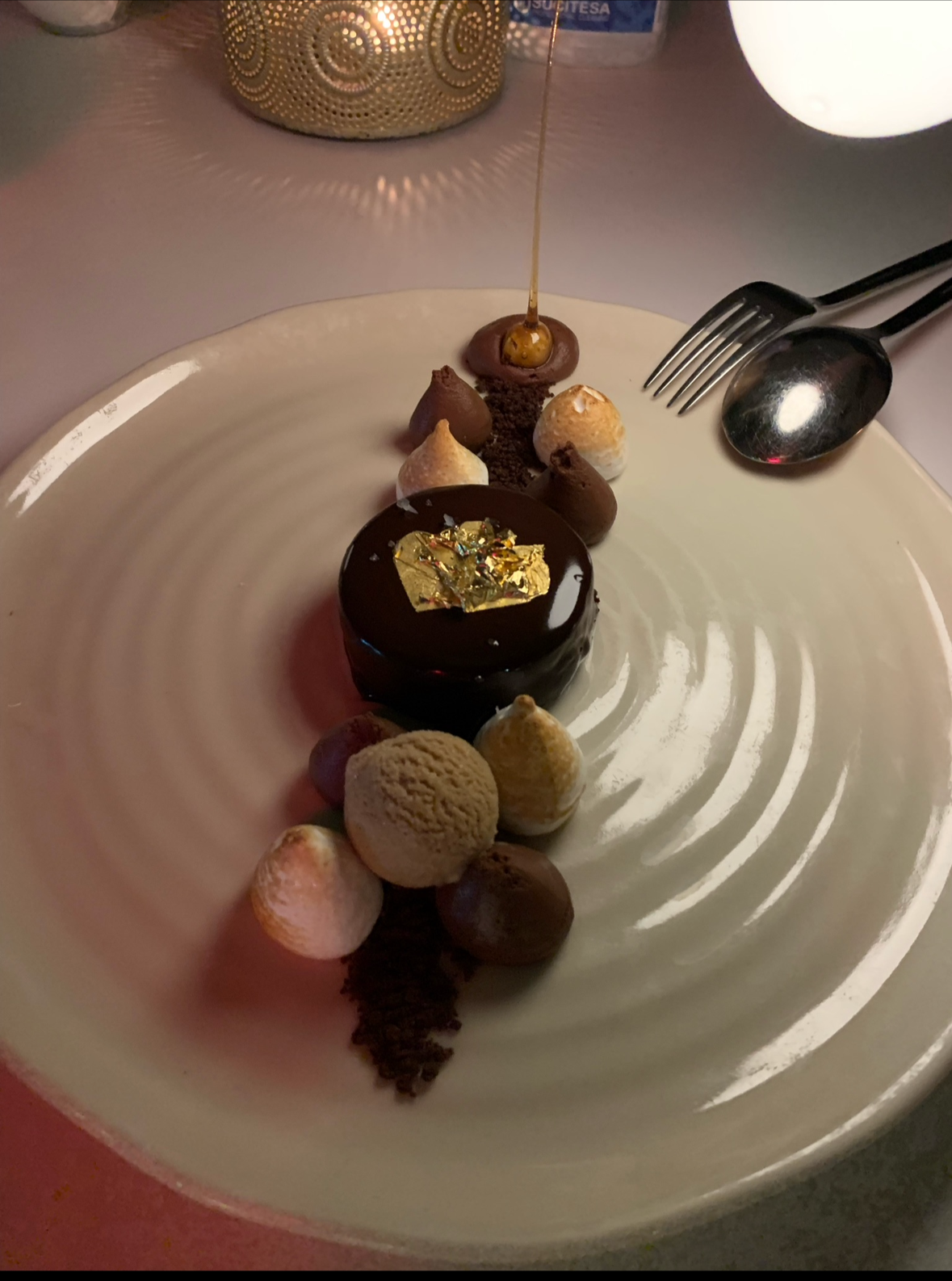 A fancy dessert plate with a small chocolate cake topped with edible gold leaf, surrounded by various flavored truffles and small meringues, with a drizzle of honey and chocolate crumbs, on a white textured plate.