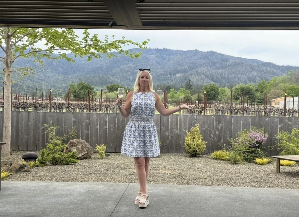 A woman in a blue and white patterned dress standing outdoors, holding a glass of wine in one hand with her other arm outstretched, with a landscape of mountains, trees, and a vineyard behind her.