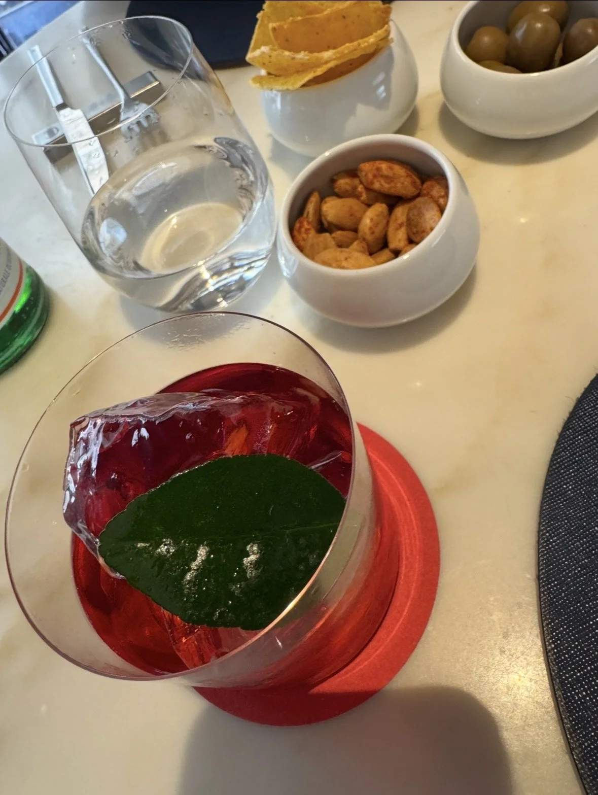 A table with various snacks and a drink, including a glass of water, a cocktail with a basil leaf, a bowl of roasted coffee beans, a bowl of green olives, and a bowl of roasted almonds, with a tortilla chip basket in the background.