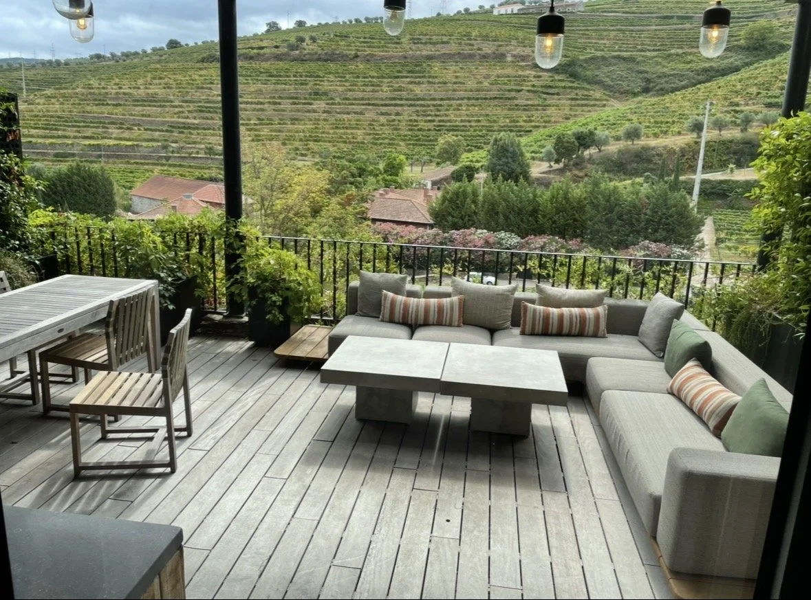 Outdoor patio overlooking a lush, terraced hillside with vineyards, featuring a sectional sofa with cushions, a rectangular coffee table, a dining table with chairs, and hanging outdoor lights.
