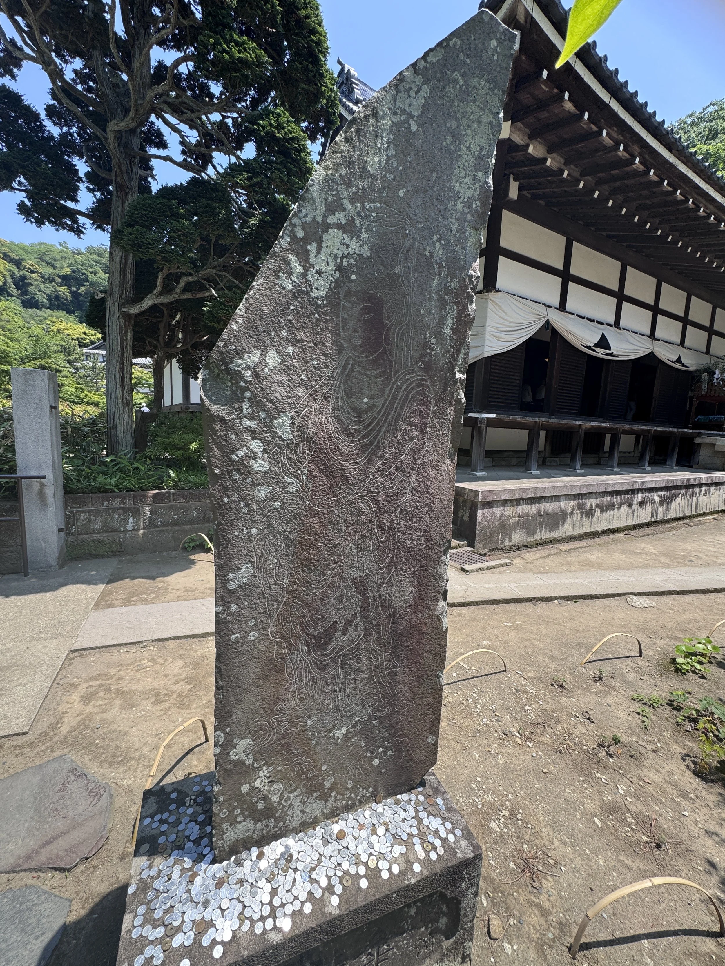A large stone with an engraved outline of a woman’s face and flowing hair stands in front of a traditional Japanese building with dark wood and white walls. The stone has lichen and weathering, and coins are scattered at its base. Tall trees and greenery are in the background.