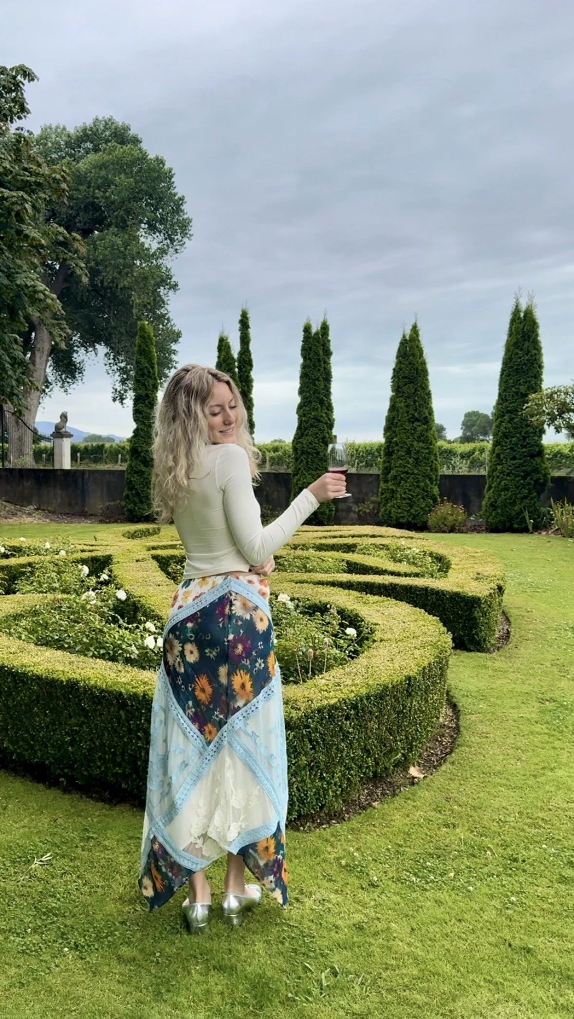 A woman with blonde curly hair smiling and holding a glass of red wine in an outdoor garden with neatly trimmed grass and tall green trees.