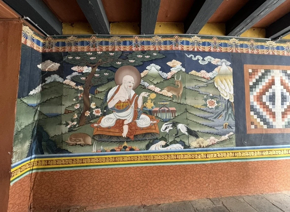 A traditional Buddha painting on a wall depicting Buddha seated outdoors, surrounded by mountains, trees, and animals, with clouds and decorative borders.