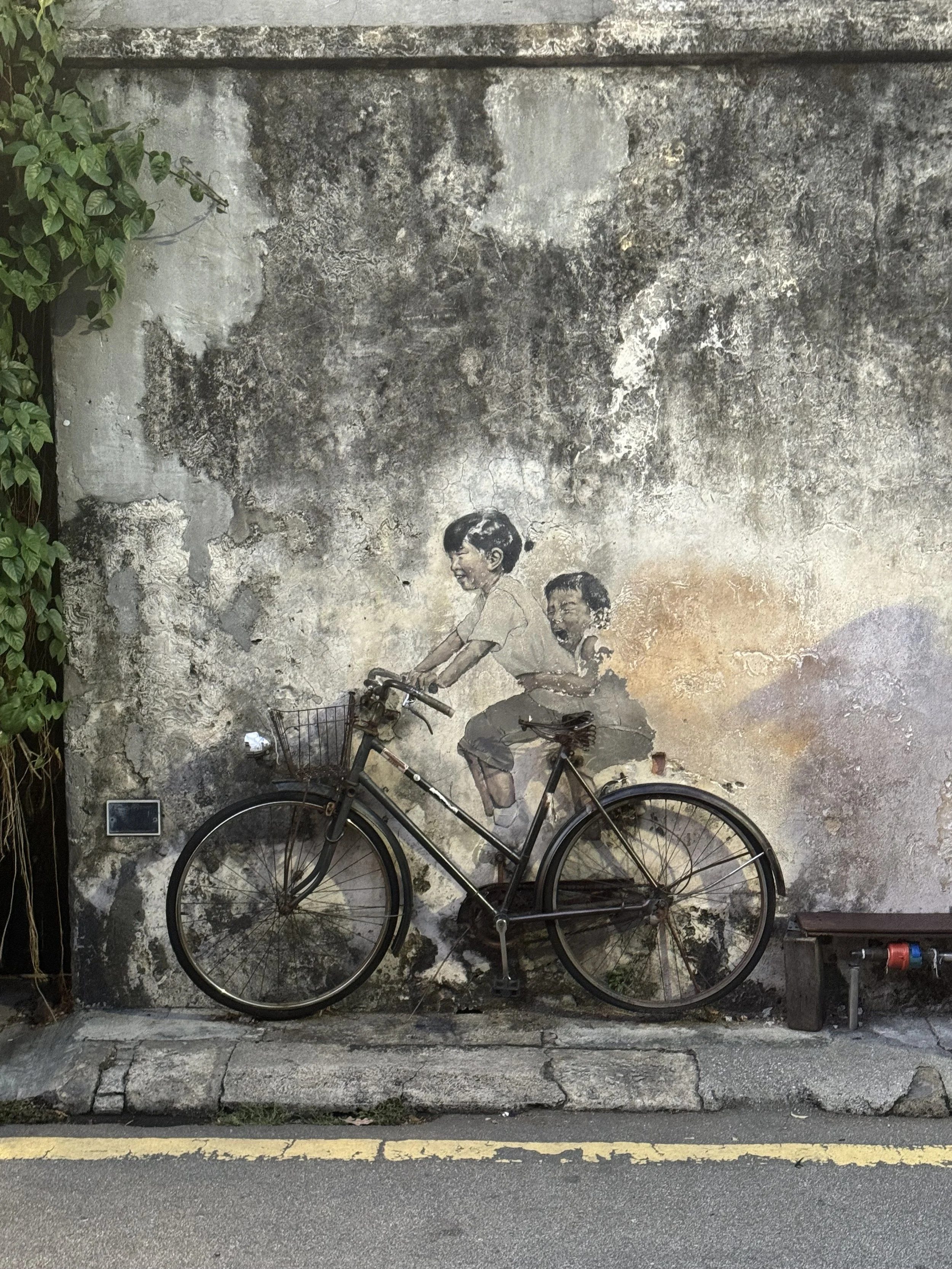 A black bicycle resting against a weathered, painted wall with a mural of two children riding a bicycle, with greenery on the left side and street curb and road with yellow lines at the bottom.