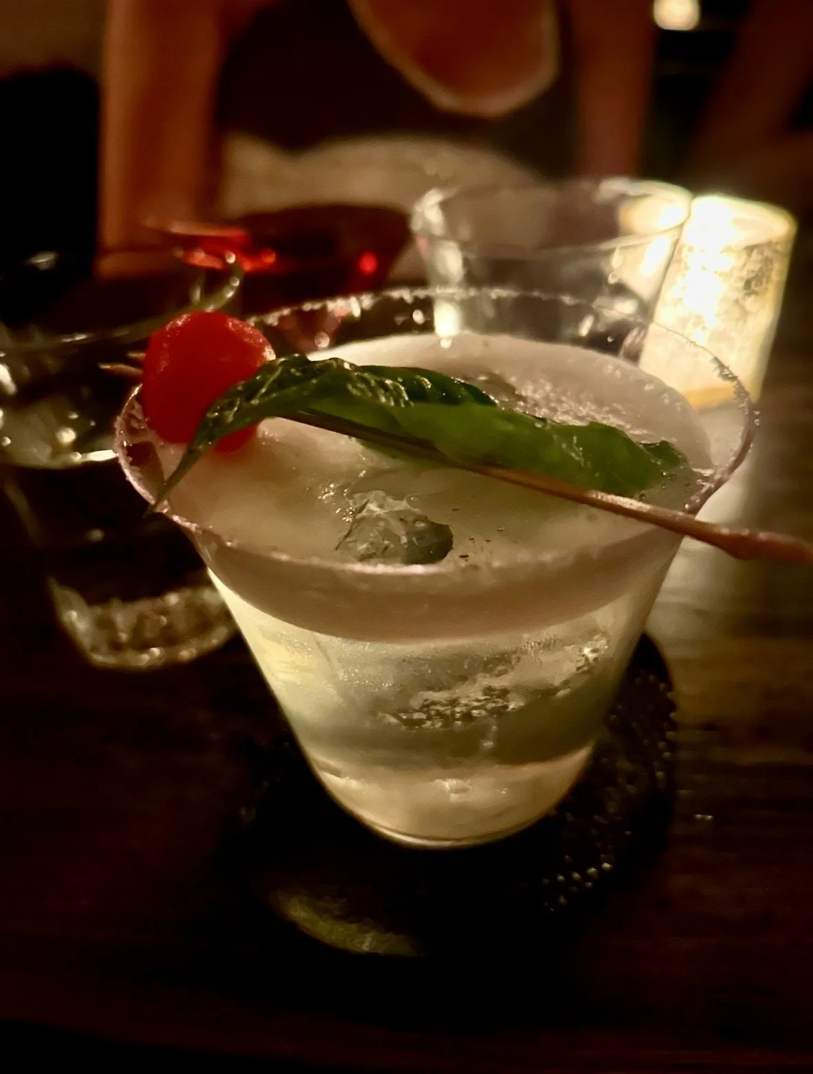 A close-up of a cocktail glass containing a clear drink with ice, garnished with a red cherry and green leaf, on a dark wooden table in a dimly lit setting.