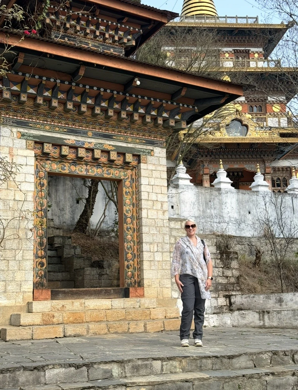 A woman standing in front of a traditional Tibetan structure with colorful, ornate decoration, a doorway without a door, and a large pagoda-style building in the background.