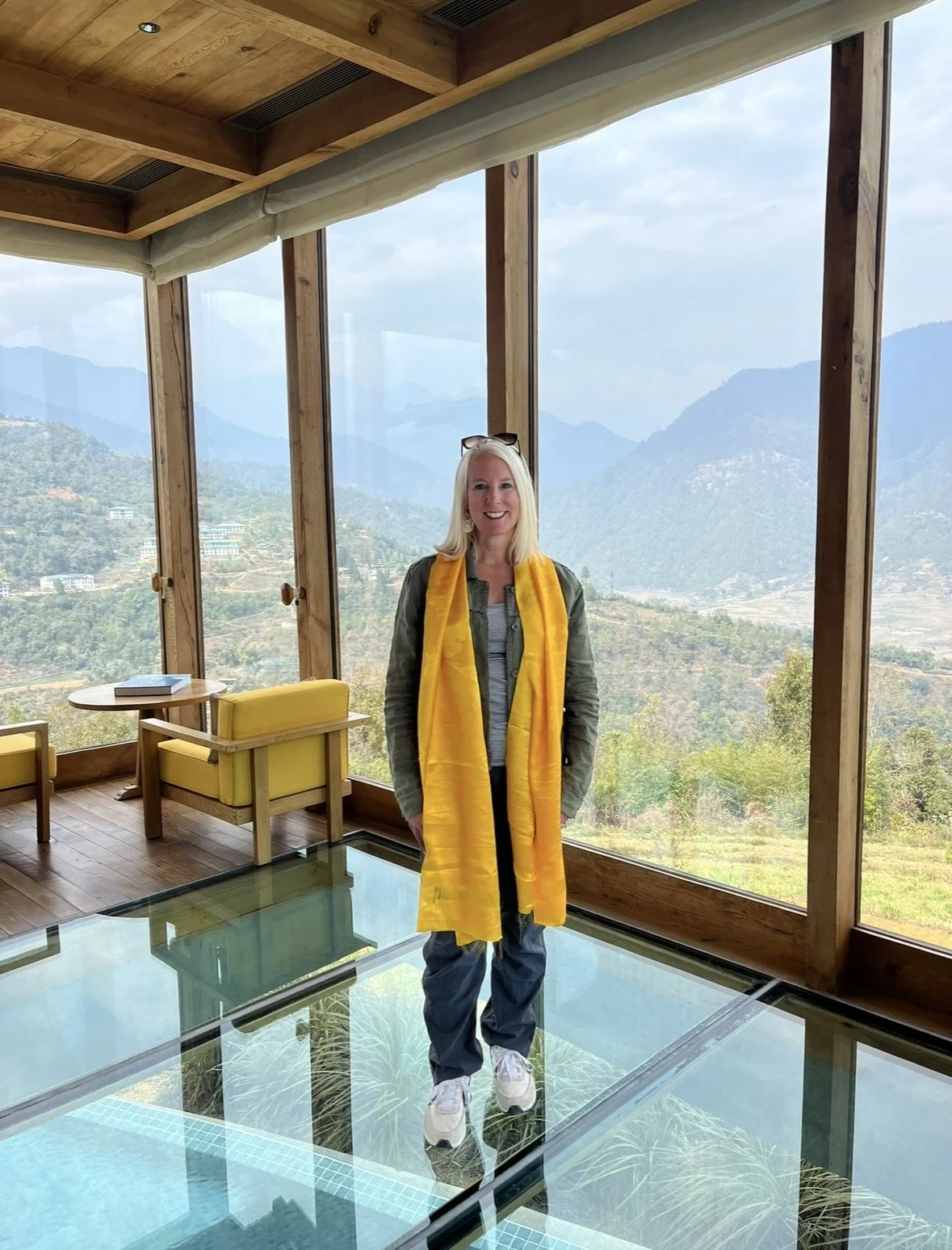A woman standing on a glass floor inside a room with large windows, with a mountainous landscape in the background, wearing a yellow scarf, gray jacket, jeans, and white sneakers.