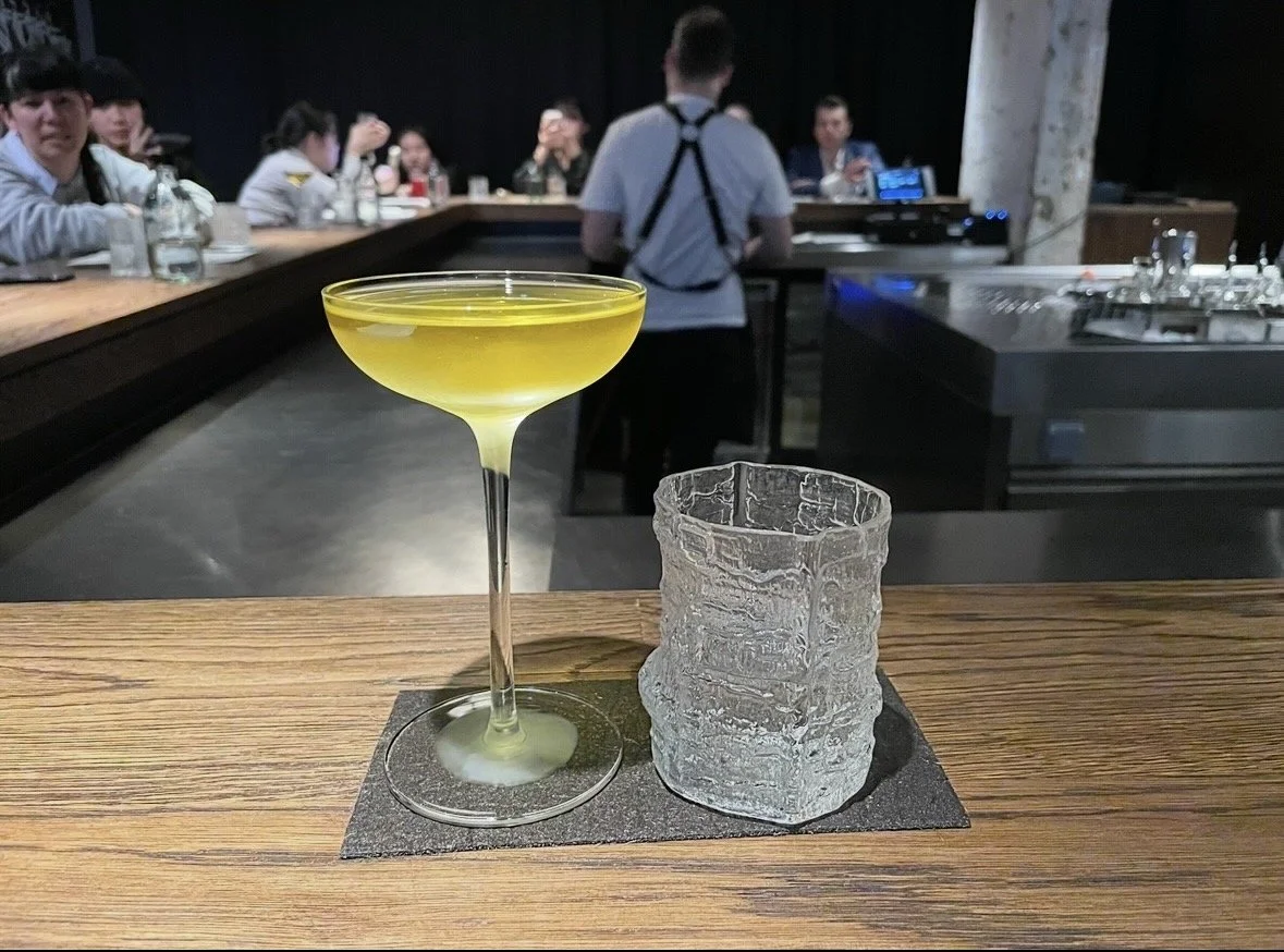 A yellow cocktail in a coupe glass next to a textured ice cube on a black napkin on a wooden bar counter.