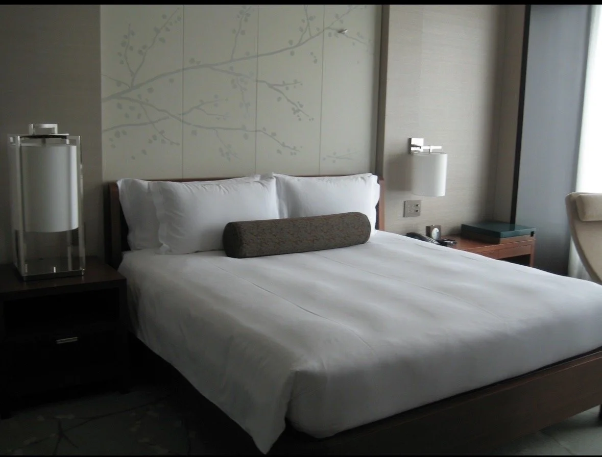 A neatly made hotel bed with white sheets, pillows, and a decorative bolster pillow in a modern hotel room. There are two bedside lamps, a nightstand with a phone, and a window with light streaming in.