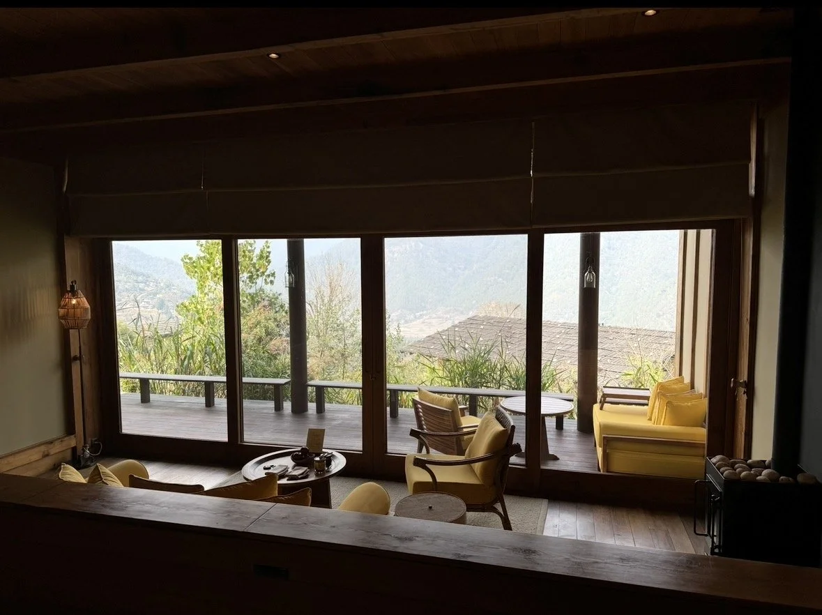 A cozy indoor seating area with yellow armchairs, a small round table, and a view of a balcony with trees and mountains through large sliding glass doors.