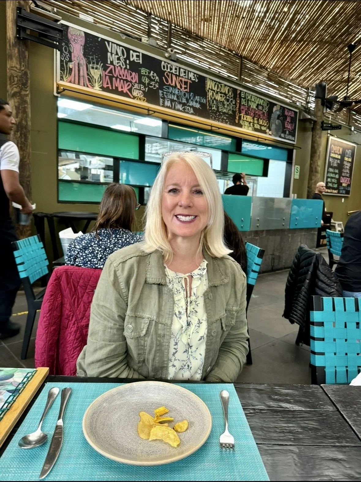A smiling woman with blonde hair sitting at a restaurant table, with a plate of potato chips and utensils in front of her. The restaurant has colorful chairs and chalkboard menus in the background.