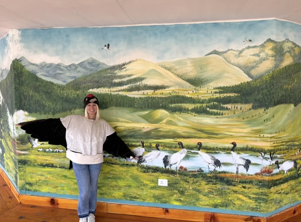 A woman smiling and dressed in a bird costume with black wings, standing in front of a large mural of a nature scene with mountains, trees, a lake, and cranes.