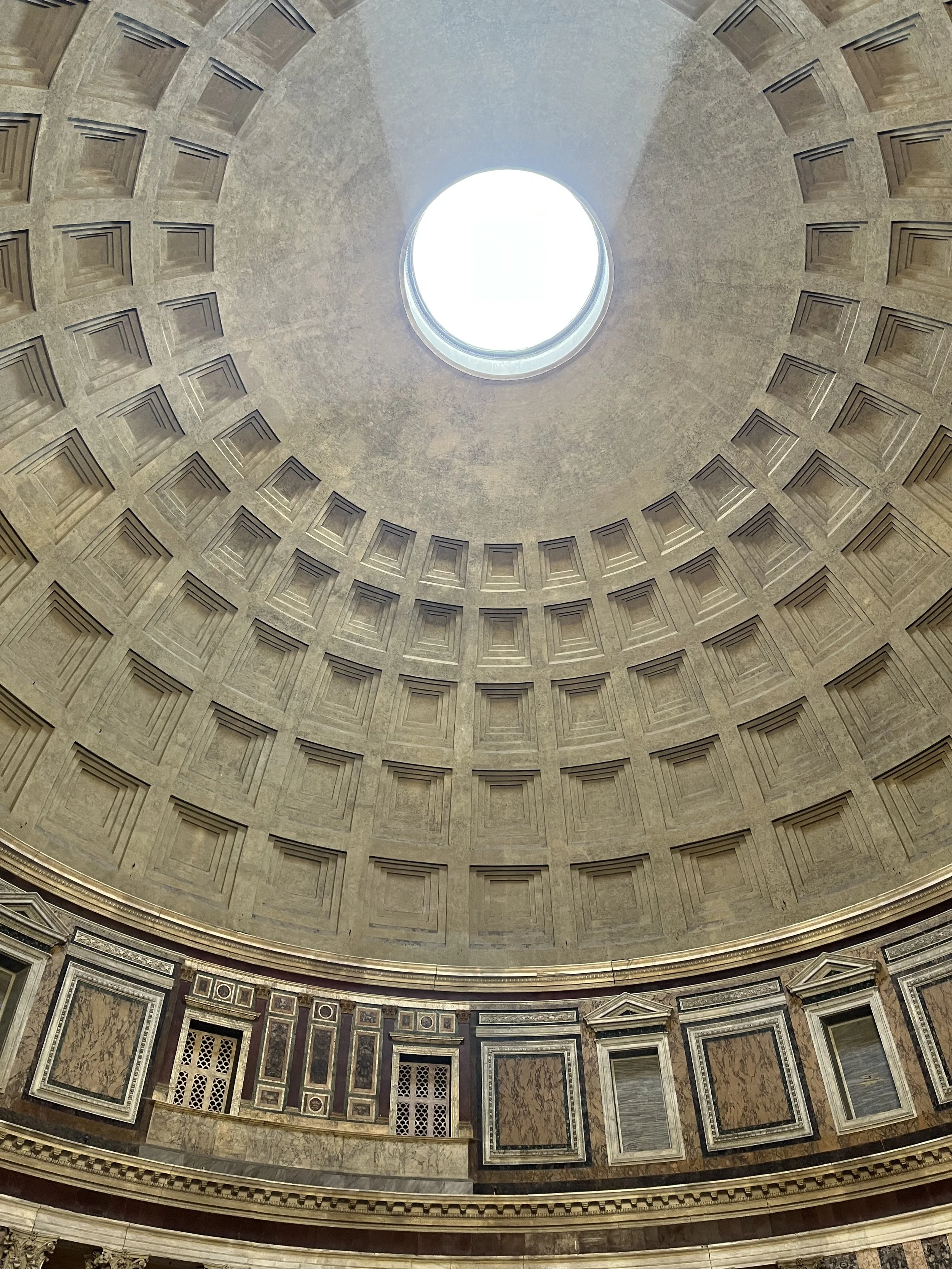 The interior of the Pantheon in Rome, with a view of the large oculus at the center of the domed ceiling, surrounded by coffered square designs.