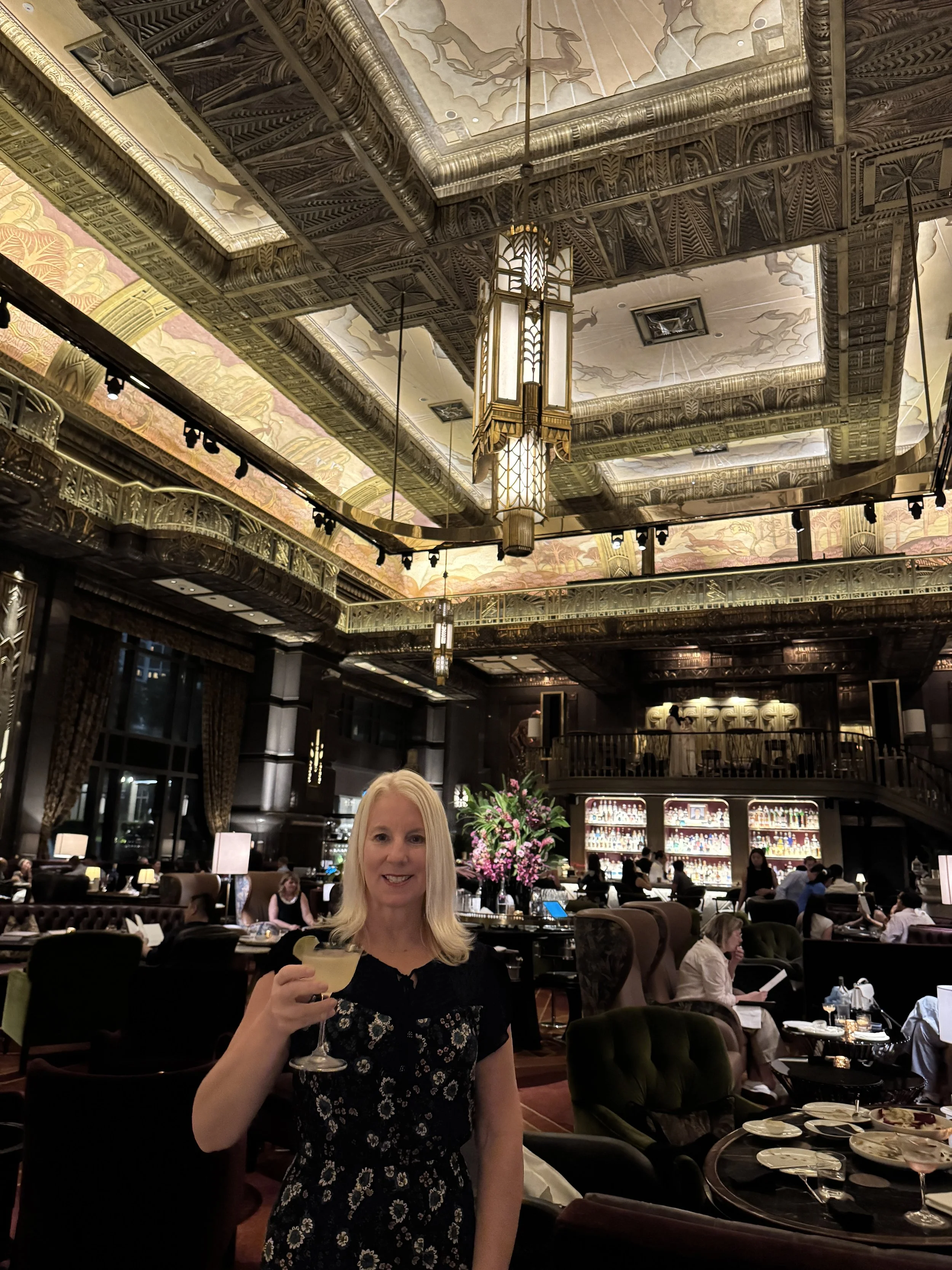A woman with blonde hair smiling and holding a glass of wine or champagne in a luxurious restaurant with ornate ceiling decor, chandeliers, and a bar in the background.