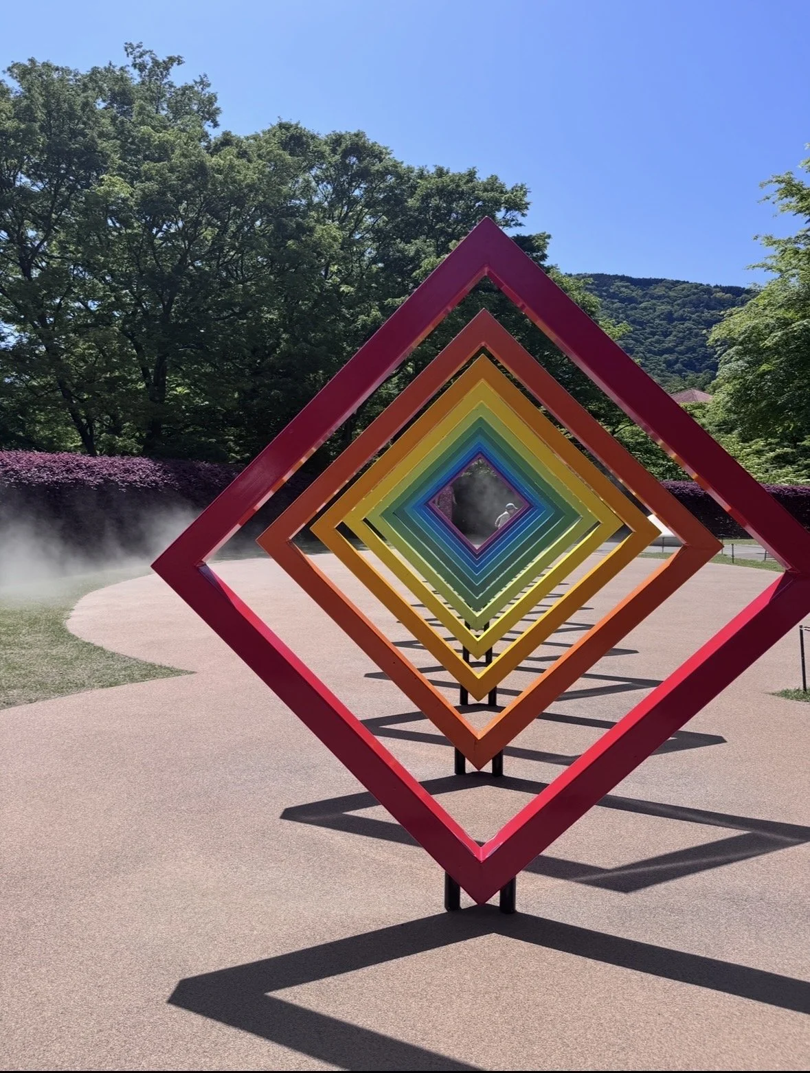 Colorful geometric outdoor sculpture consisting of concentric diamond frames in red, orange, yellow, green, blue, and purple, creating a tunnel effect in a park setting with trees and a mountain in the background on a sunny day.