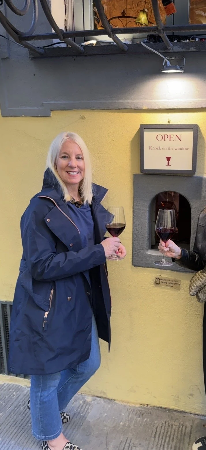 A smiling woman with long blonde hair in a blue jacket and jeans holding a glass of red wine outside a building with a yellow wall. There is a sign that says 'OPEN. Knock on the window,' and another person partially visible holding a similar glass of red wine.