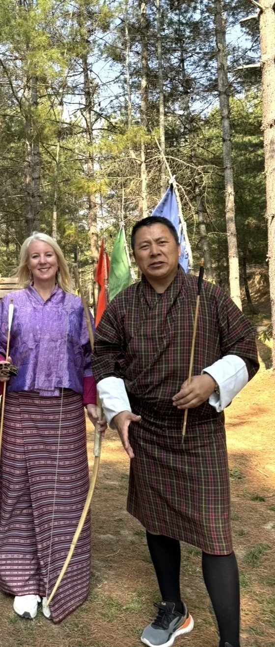 Two people dressed in traditional clothing standing in a forest, holding bows and arrows.