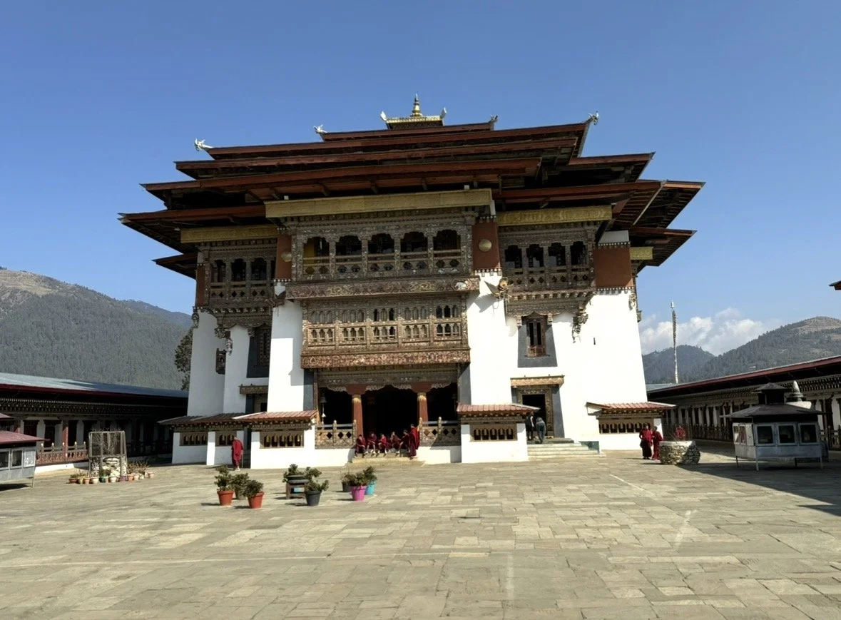 Traditional Bhutanese monastery with intricate woodwork, set against a mountainous landscape and a clear blue sky.