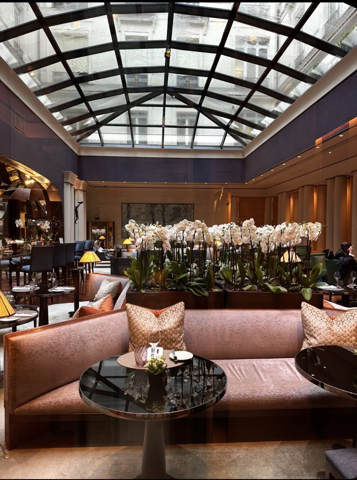 Elegant hotel lobby with a large glass ceiling, arranged seating with pillows, a large flower display with white orchids, and dim lighting.