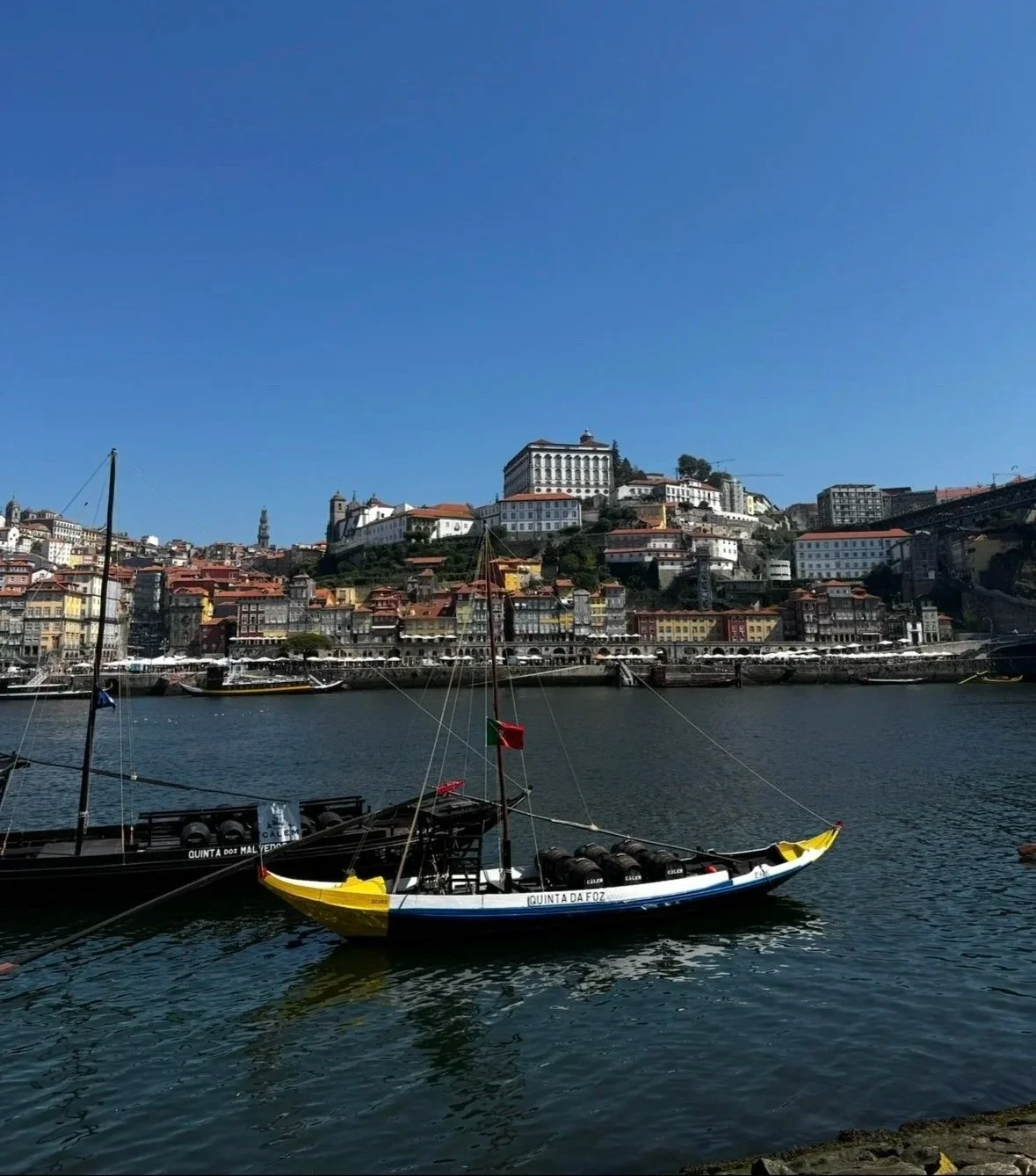 Boats on a river with a hillside cityscape in the background, featuring colorful buildings and historic architecture under a clear blue sky.