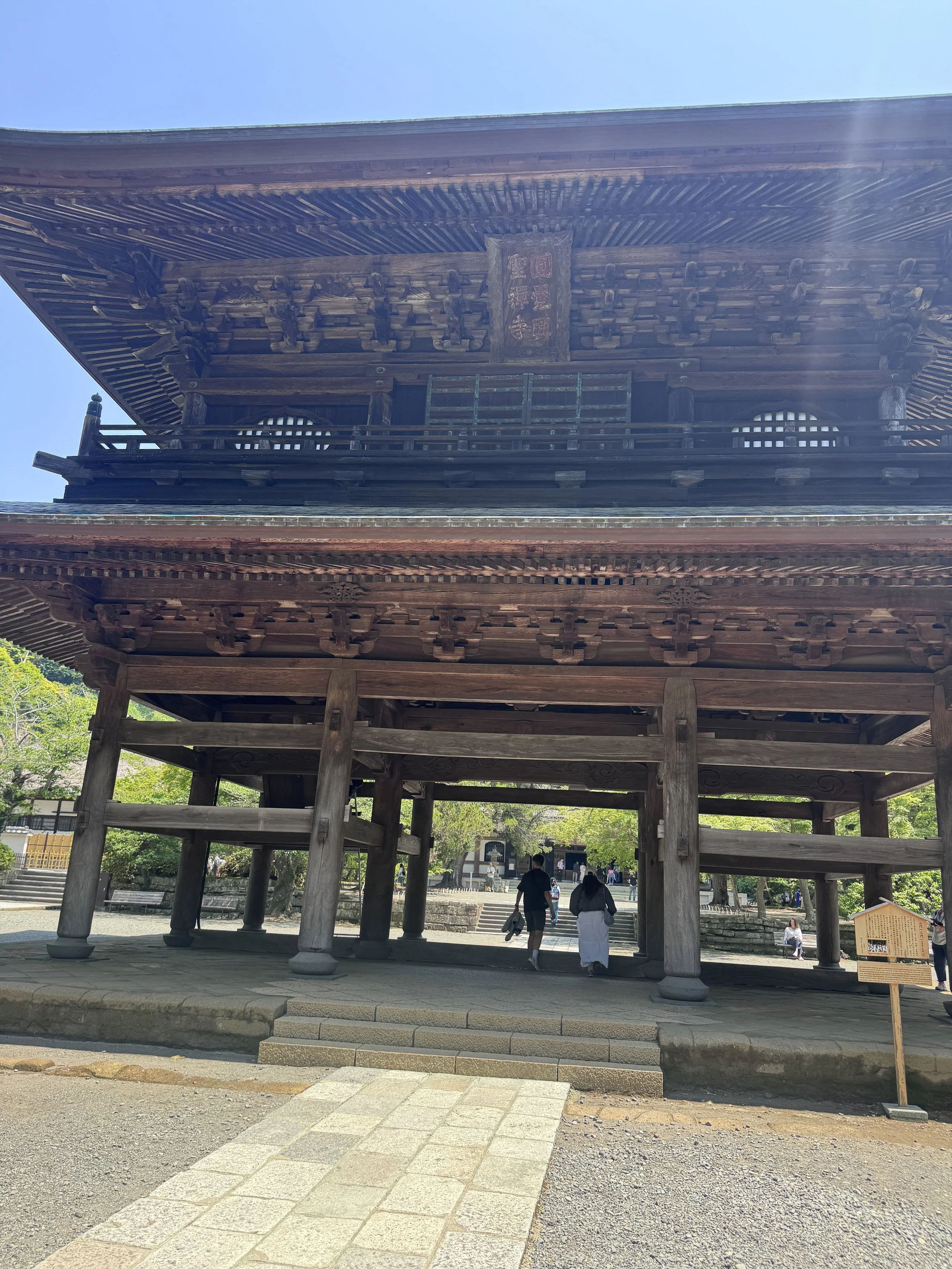 A traditional Japanese wooden temple with multiple tiers, intricate wooden detailing, and a raised platform accessed by stairs, situated outdoors on a sunny day with a few people walking beneath it.