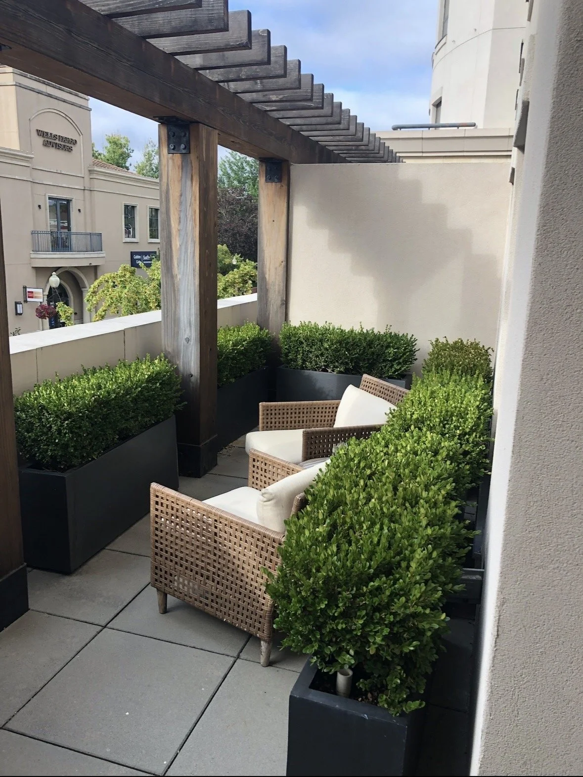 Small outdoor balcony with two wicker armchairs with white cushions, surrounded by green potted bushes and a wooden pergola on top.