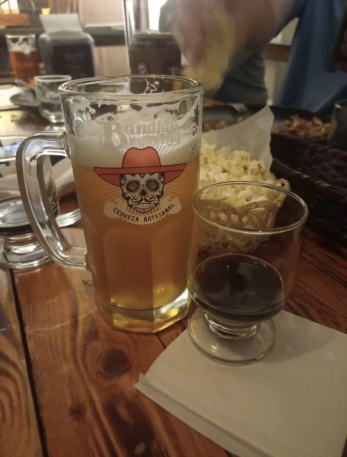 A mug of beer with a skull and sombrero logo, a small glass of red wine, a bowl of popcorn, and a plate of food on a wooden table.