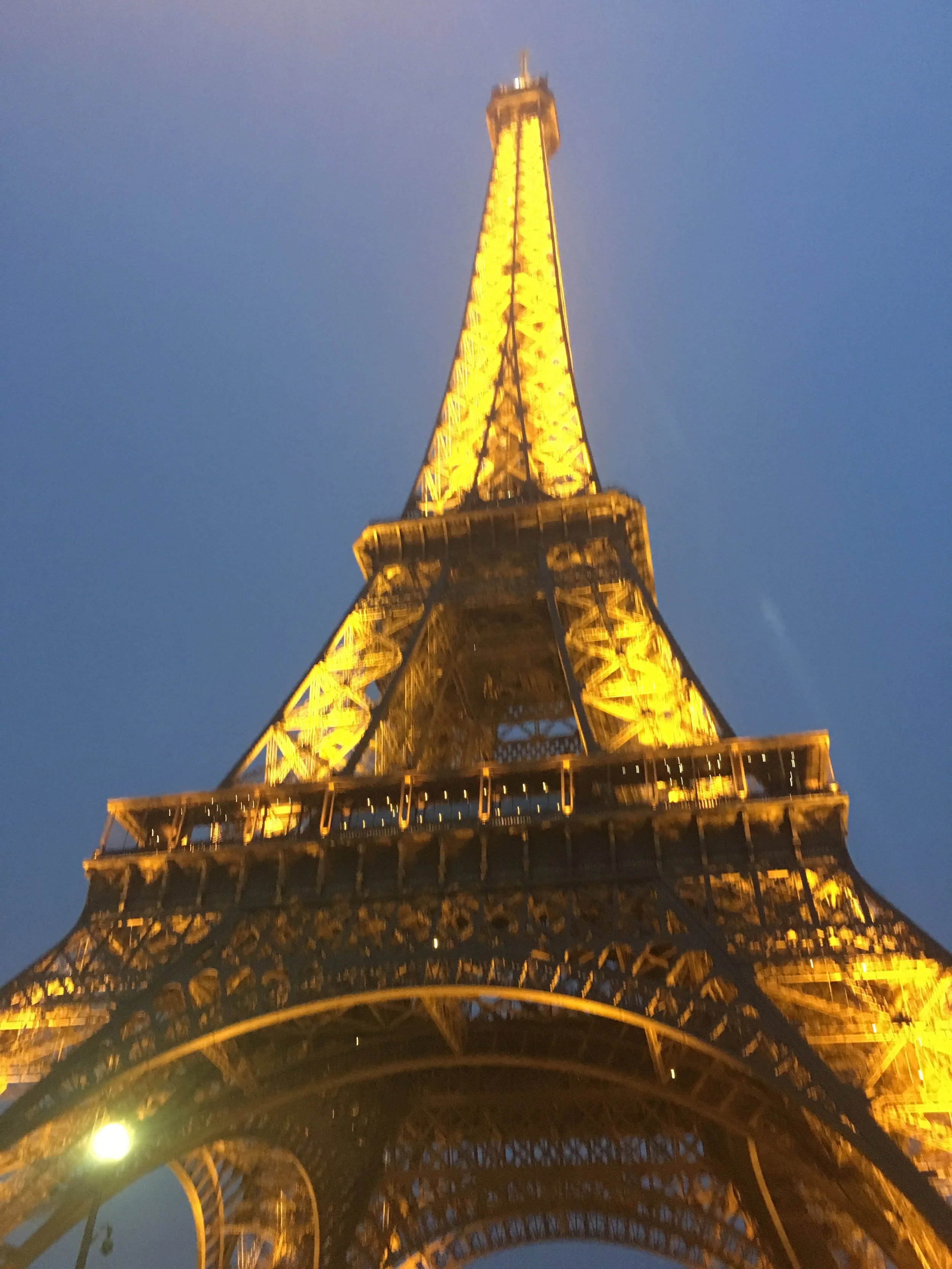 The Eiffel Tower illuminated with yellow lights at night, viewed from beneath the structure against a dark blue evening sky.