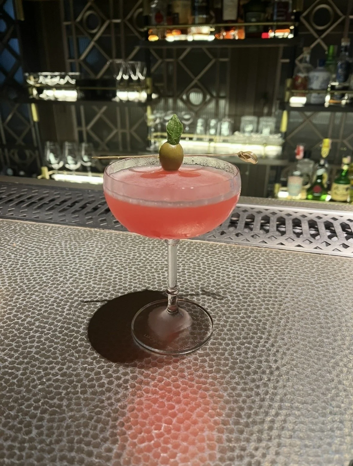 A pink cocktail in a coupe glass garnished with a green olive and a small leaf, sitting on a bar counter with a blurred bar background.