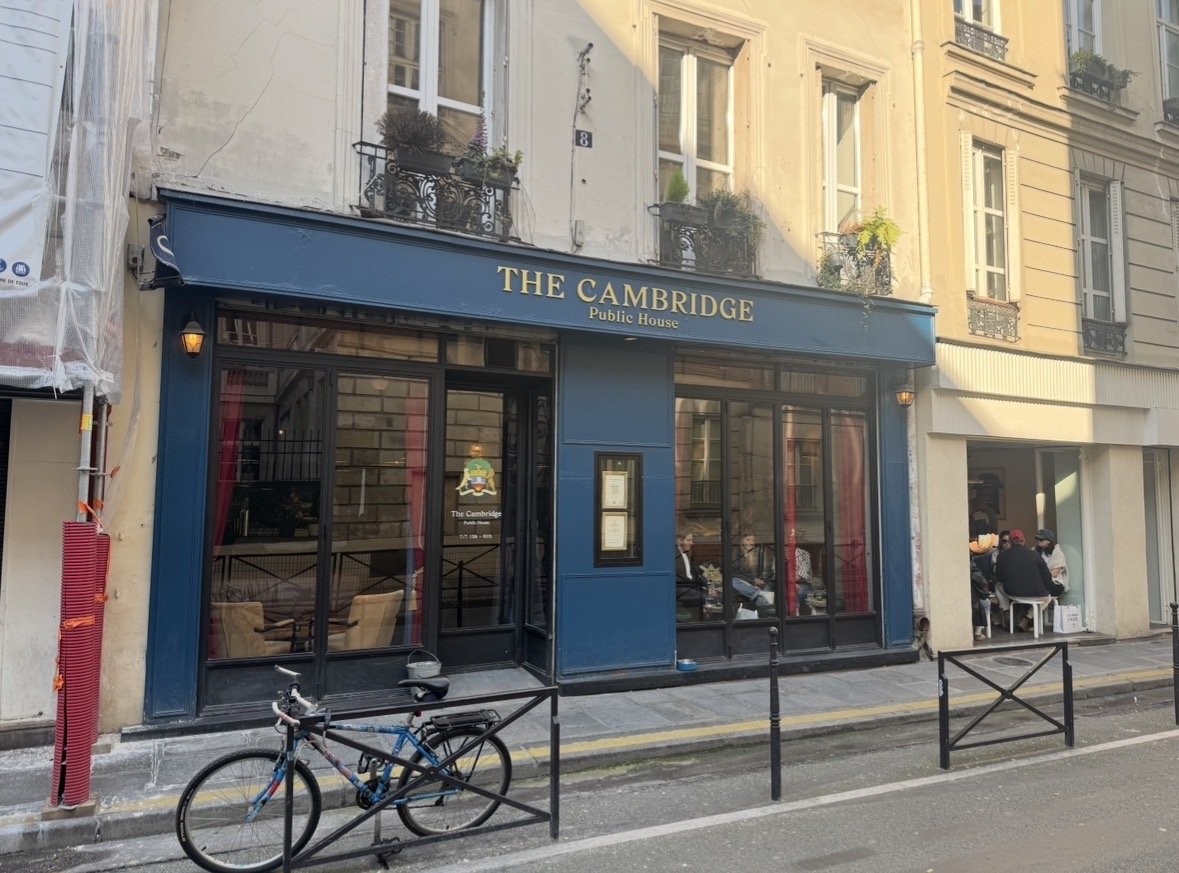 Exterior of The Cambridge Public House, a pub on a city street with large glass windows, a bicycle parked outside, and people sitting at tables on the sidewalk.