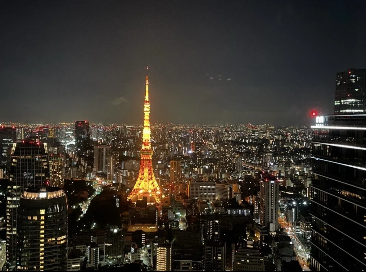 Nighttime cityscape of Tokyo, Japan, featuring the illuminated Tokyo Tower in the center with surrounding high-rise buildings and a dark sky.