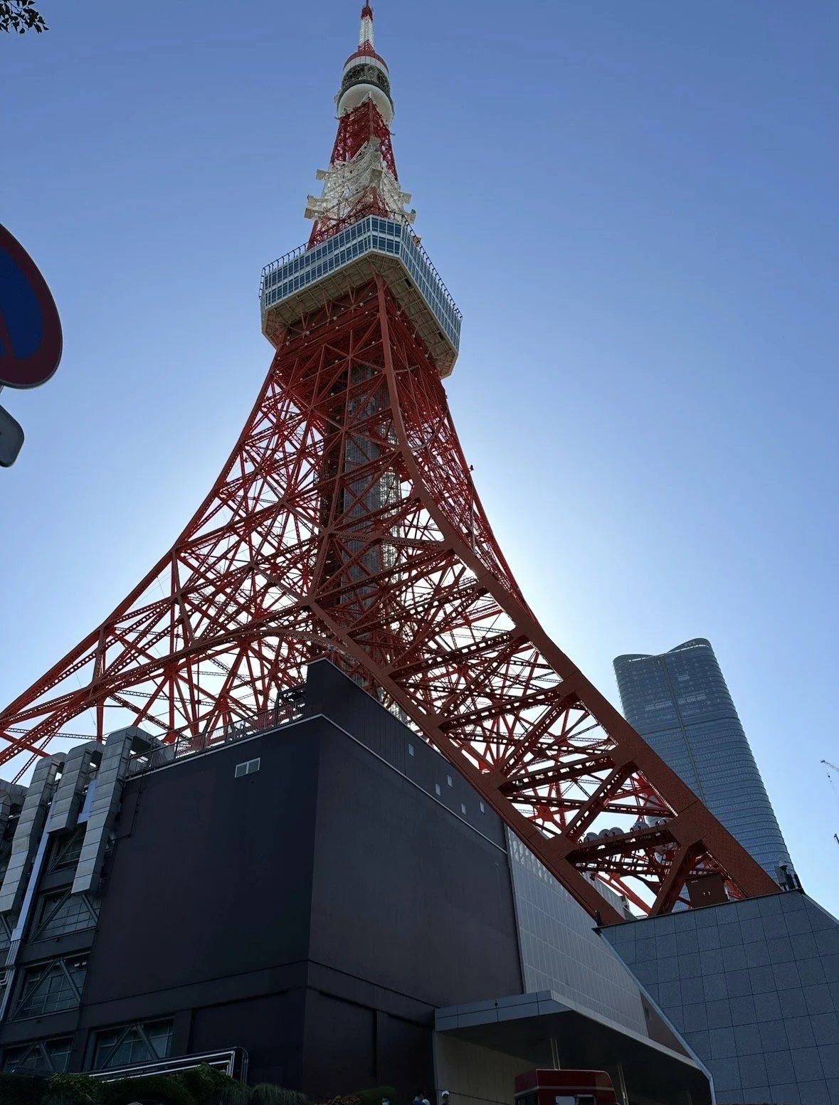 A tall red and white communication tower with a viewing platform and antenna, set against a blue sky, with modern buildings in the background.