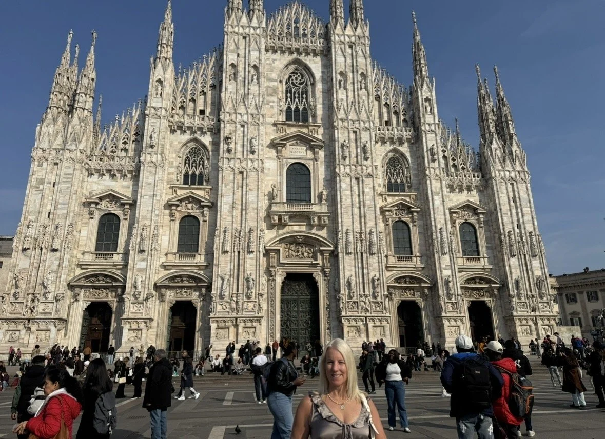 A woman standing in front of the Milan Cathedral, also known as Duomo di Milano, a large Gothic church with intricate white marble facade and multiple spires, during daytime with a crowd of tourists.