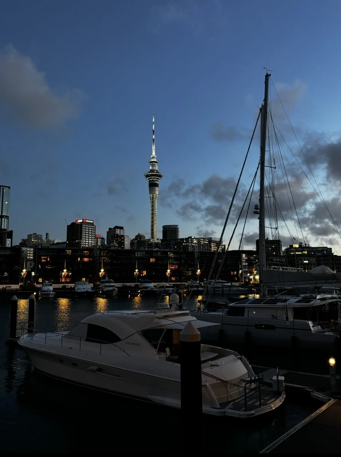 A marina with boats docked at sunset, city skyline with a tall tower in the background, and a cloudy sky.