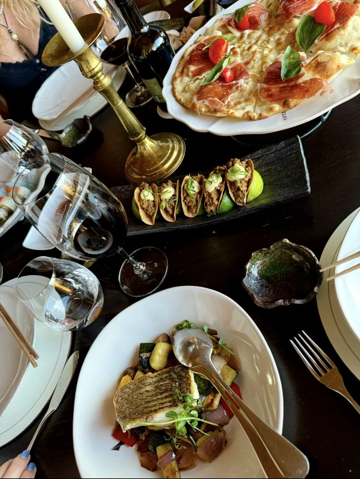 A table with various dishes including a pizza with prosciutto and basil, a plate of grilled fish with vegetables, small tacos with green garnish on a black wooden serving board, and several glasses of red wine. A gold candlestick with a white candle is also on the table.