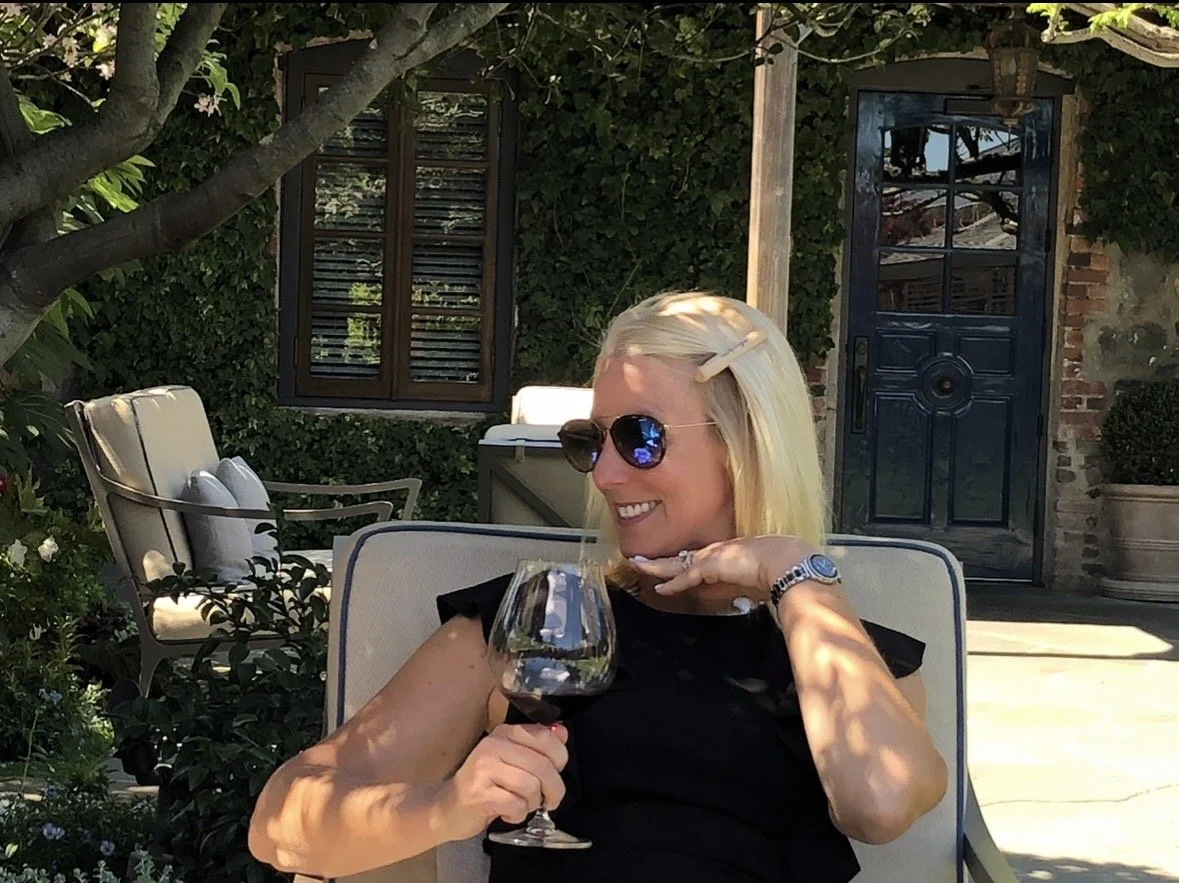 A woman with blonde hair wearing sunglasses, smiling, and holding a glass of red wine in an outdoor garden setting with green foliage, a tree, and a blue door in the background.