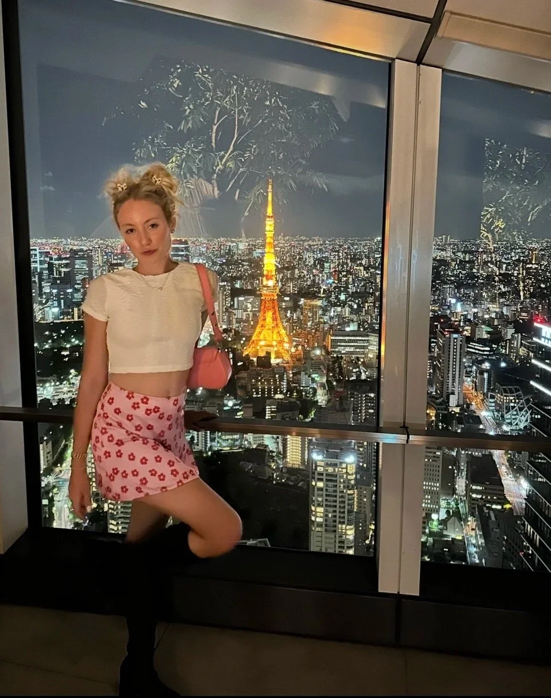 A young woman with blonde hair styled in buns, wearing a white crop top and a pink skirt with red flowers, stands in front of a large window at night. The cityscape behind her features the brightly lit Tokyo Tower, with numerous buildings and city lights extending to the horizon.