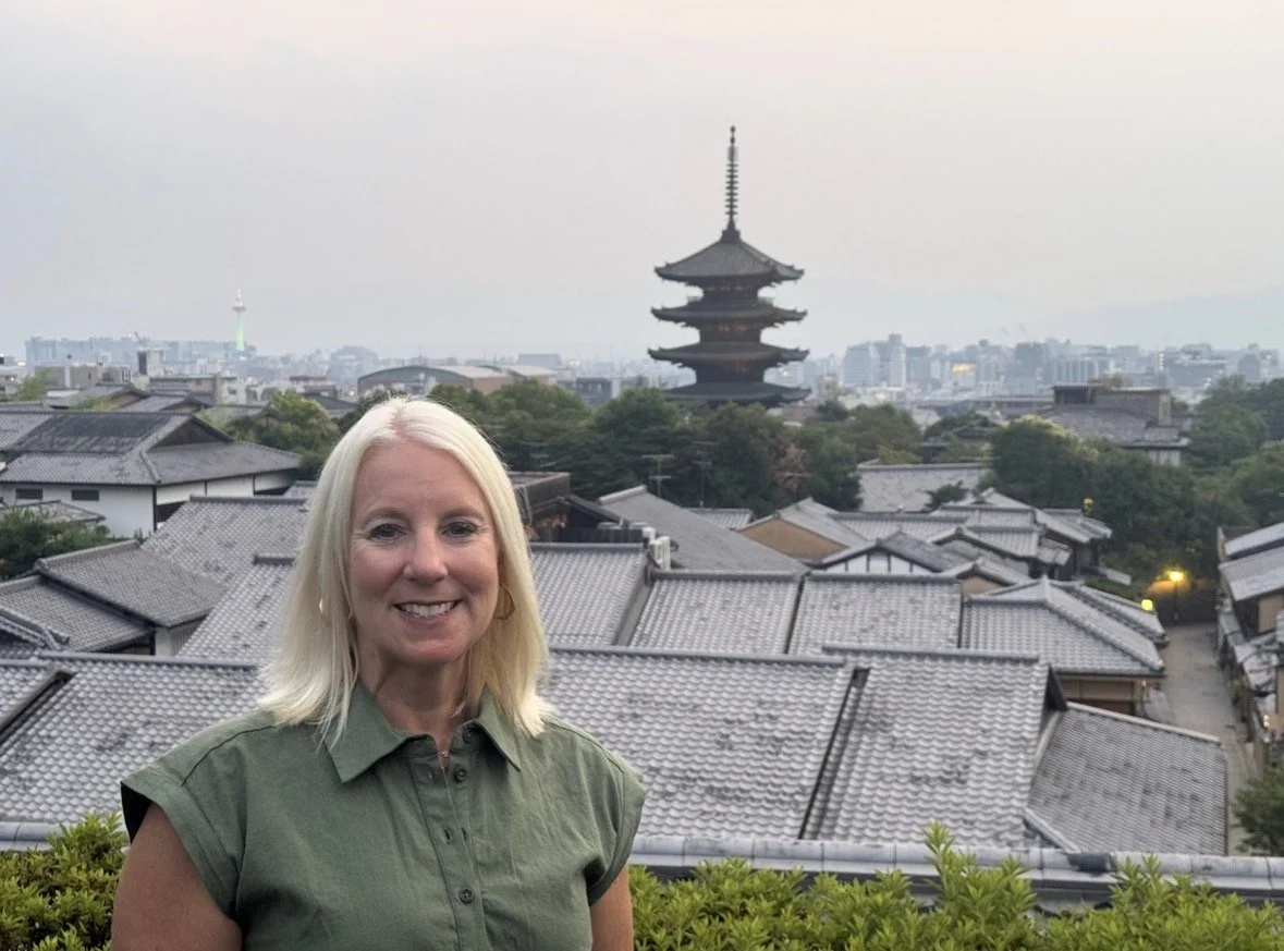 Woman with blonde hair smiling in front of traditional Japanese rooftops and a pagoda amidst greenery.