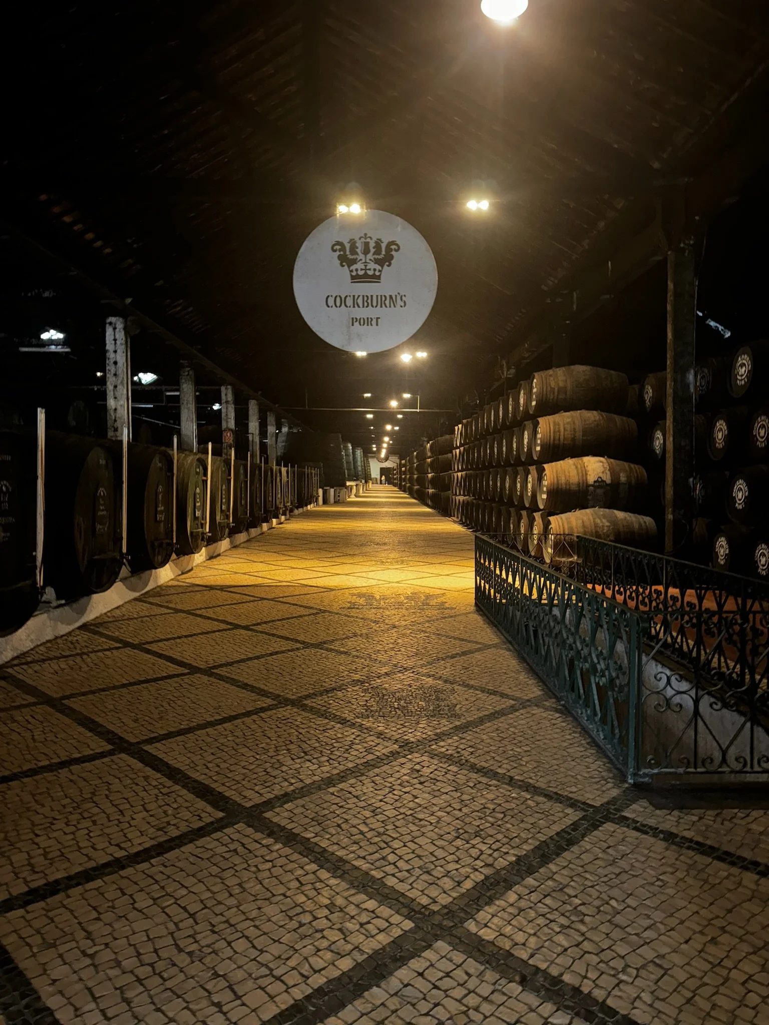 The image shows an underground cellar or storage area with stacked barrels. A sign hanging from the ceiling reads "Cockburn's Port" and features a crown symbol. The area is illuminated by ceiling lights and has a tiled mosaic floor with a diamond pattern.