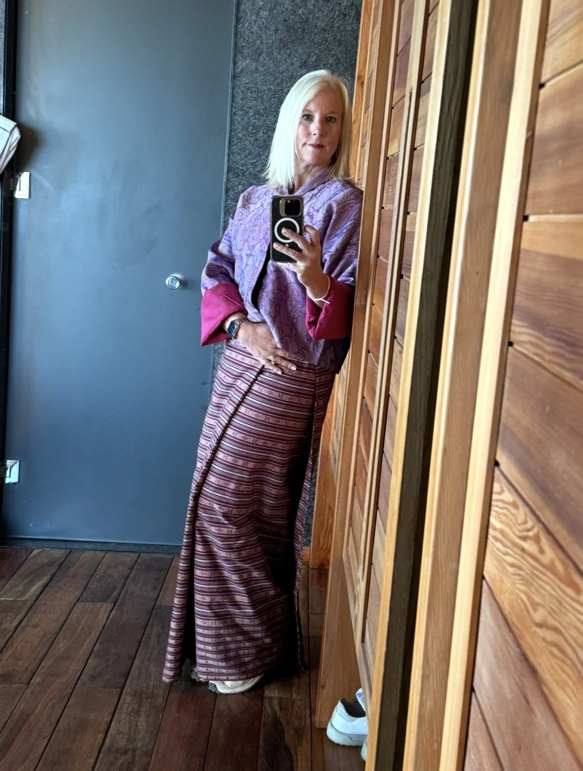 A woman taking a mirror selfie in a room with wooden walls. She is wearing a purple patterned top with red cuffs, a striped skirt, and white shoes visible at the bottom.