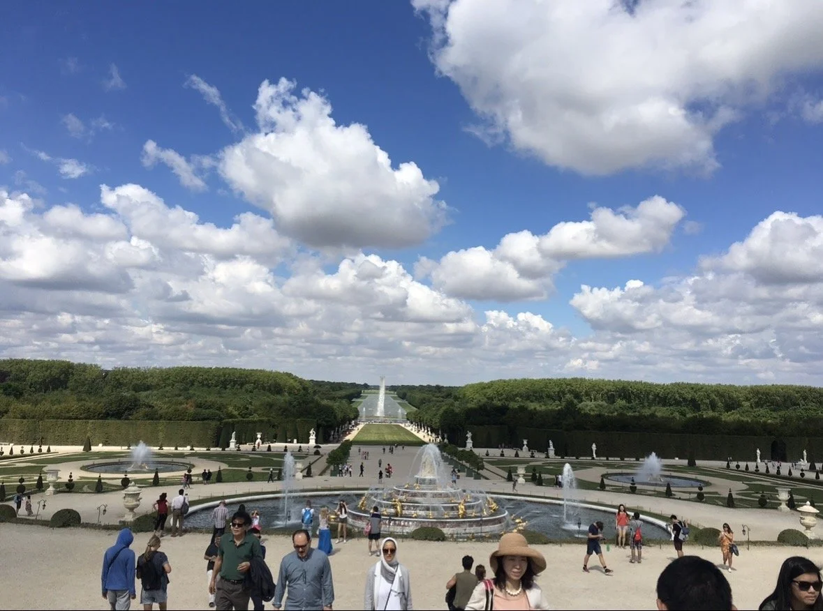 View of a large formal garden with fountains, walkways, and green hedges, with many visitors walking around on a sunny day with scattered clouds.