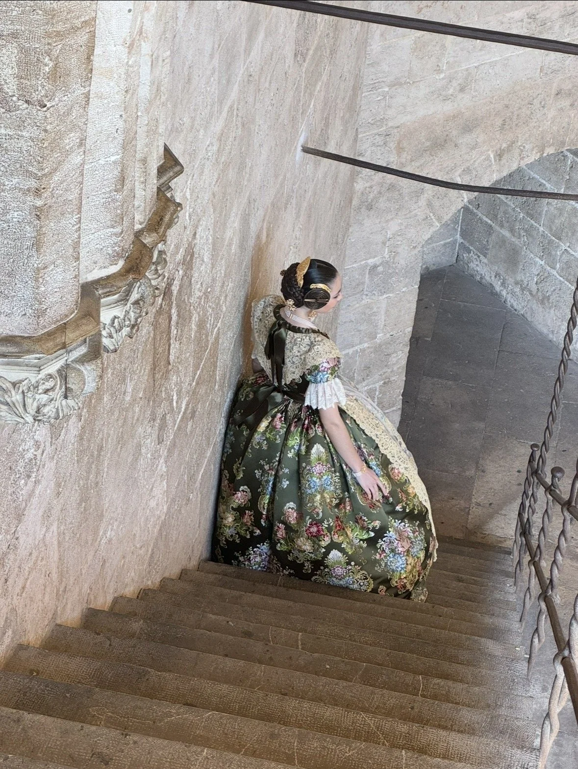 A woman dressed in traditional, ornate clothing, standing on a stone staircase in what appears to be an old stone building.